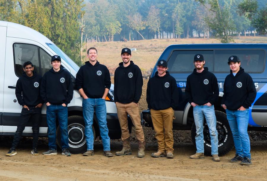 Seven team members of better basement & waterproofing company standing in front of company van, truck, and trailer.
