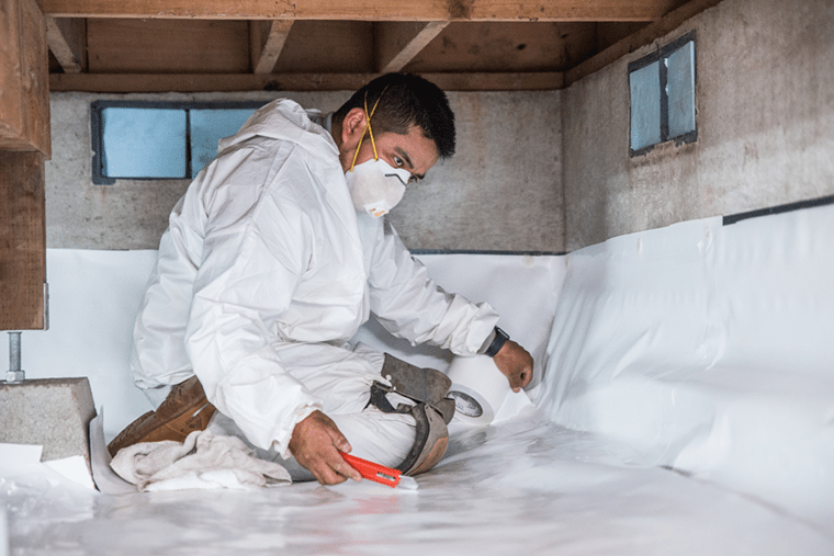 Male worker dressed in a white coverall suite in a crawl space under a house with a white PVC vapor barrier on the ground.